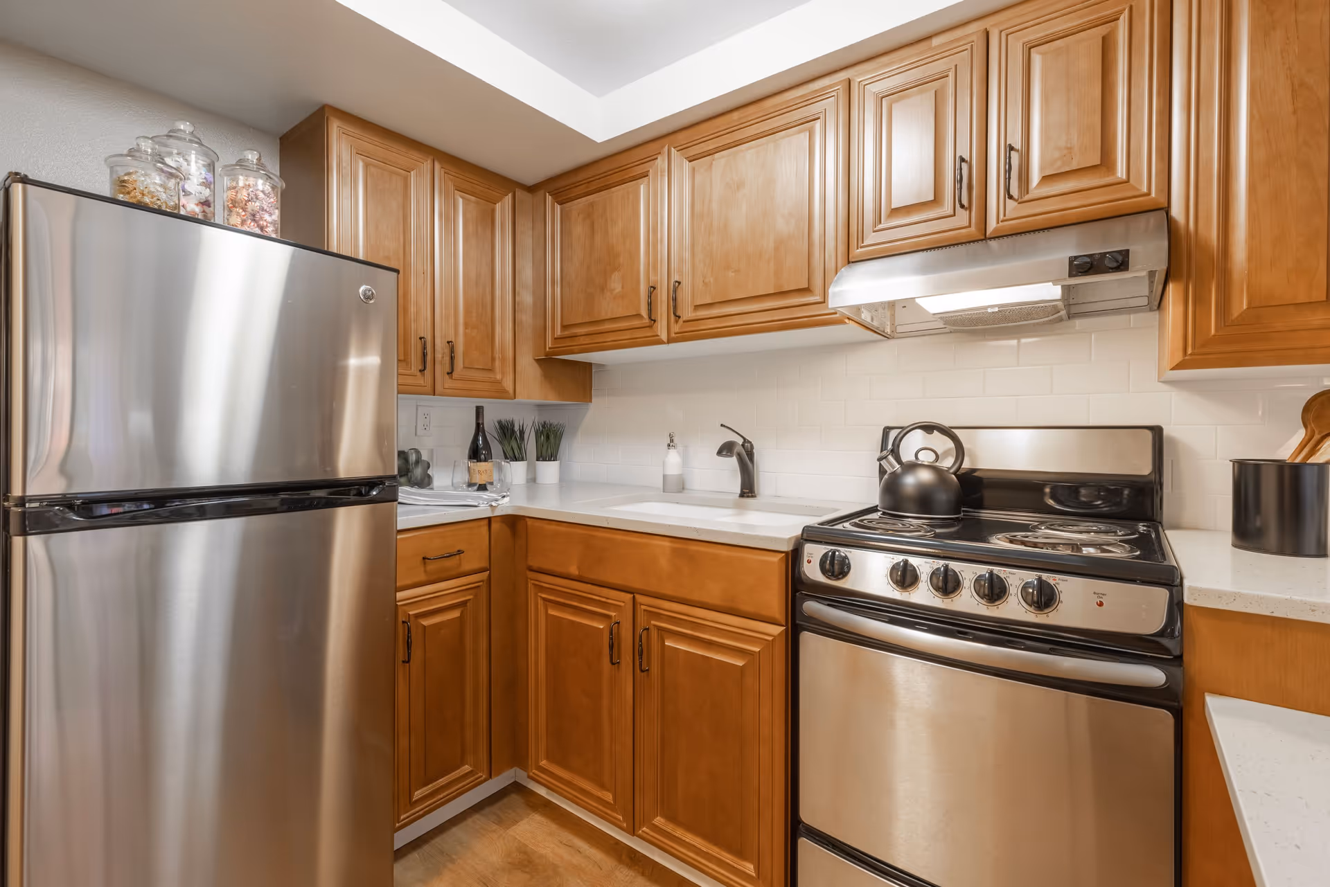 A kitchen with wooden cabinets, a stainless steel refrigerator, a stove with a kettle on it, a white countertop with a sink, and some decorative items including jars with snacks and small potted plants.