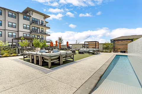 Outdoor courtyard at Sage Plum Creek with cushioned lounge seating, cabanas, umbrellas, a narrow pool, and a multi-story building under a blue sky.