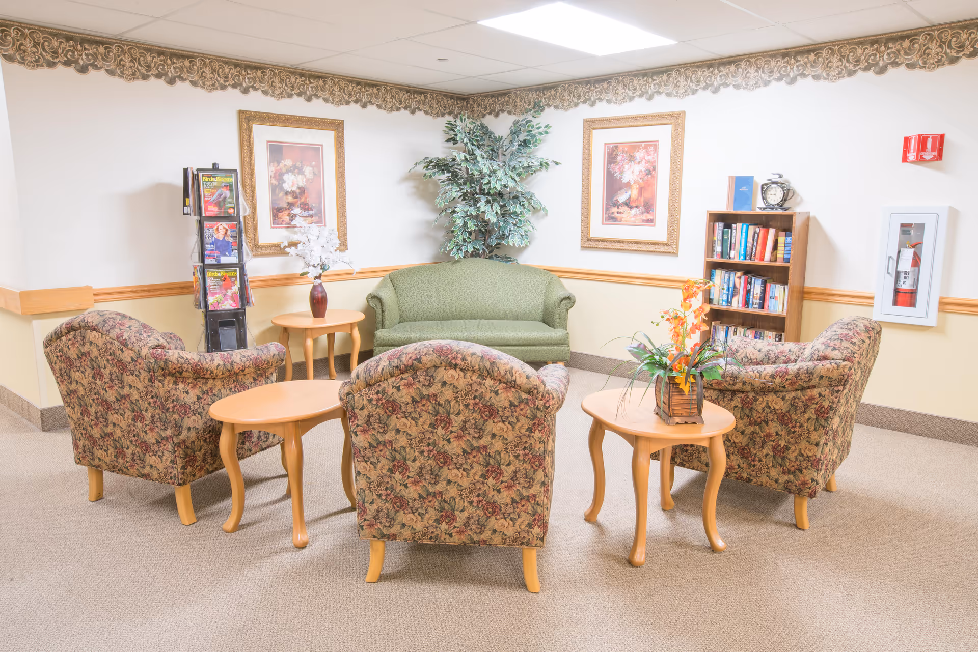A cozy sitting area in a senior living facility with a green upholstered sofa, three floral patterned armchairs, and three wooden side tables. The walls are decorated with two framed floral paintings, a tall artificial plant, a bookshelf filled with books, a fire extinguisher in a wall case, and a magazine rack.