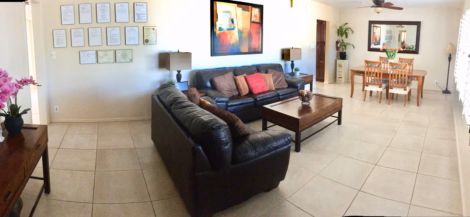 A spacious, well-lit living and dining area with beige tiled floors. The living room features two black leather sofas arranged around a wooden coffee table with decorative items. The dining area has a wooden table with six chairs and a large mirror on the wall. The walls are decorated with framed certificates and a colorful abstract painting. There are two table lamps on side tables, a potted plant, and a vase with flowers on a side table near the entrance.