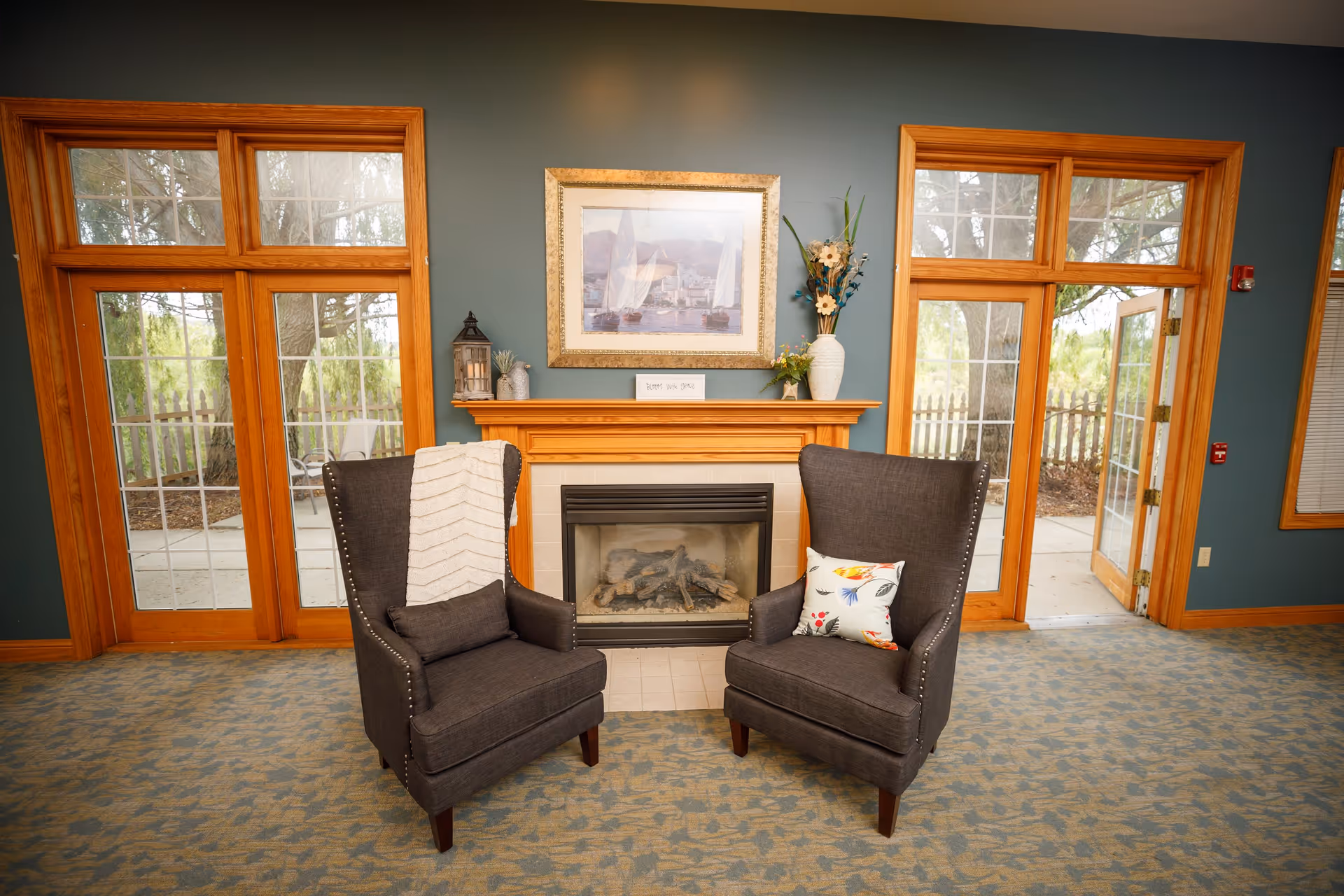 A cozy sitting area with two dark gray armchairs placed in front of a fireplace. One chair has a white throw blanket draped over it, and the other has a decorative pillow with a floral pattern. The fireplace mantel is decorated with a framed picture of sailboats, a lantern, and vases with flowers. Large wooden-framed glass doors and windows surround the area, letting in natural light and showing an outdoor patio and greenery.