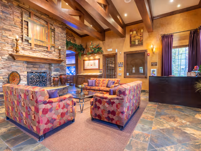 A cozy senior living facility common area with patterned sofas arranged around a glass coffee table on a rug. The room features a stone fireplace with a wooden mantel, exposed wooden ceiling beams, warm-toned walls, and tiled flooring. There is a reception desk to the right with a window behind it, and decorative plants and artwork adorn the space.