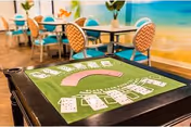 Green felt card table with playing cards in the foreground and dining tables and colorful chairs in a bright common area with a beach mural.