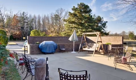 Outdoor patio area with metal chairs, a covered grill, a striped umbrella, and a wooden swing beside a low brick wall and trees.
