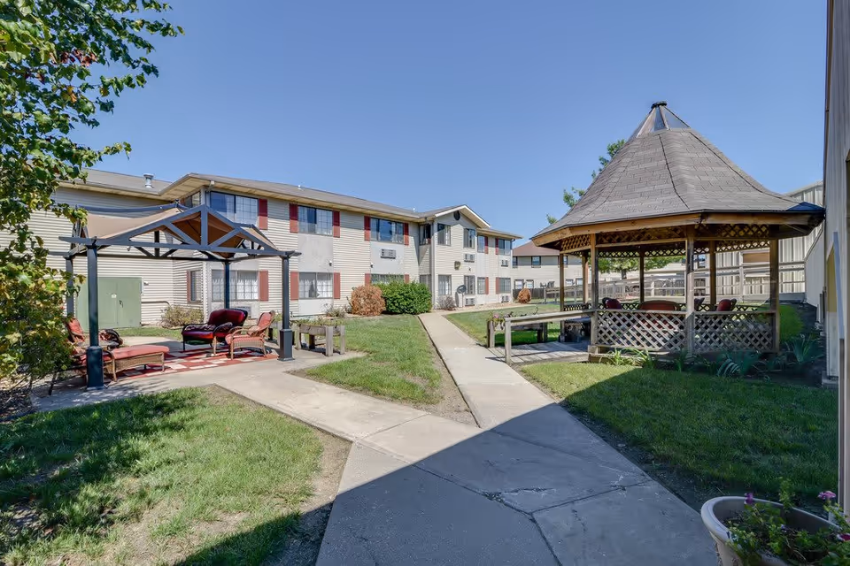 Outdoor courtyard area of a senior living facility with a gazebo on the right and a pergola with seating on the left. The courtyard is surrounded by a two-story building with beige siding and red shutters. There are paved walkways and green grass areas under a clear blue sky.