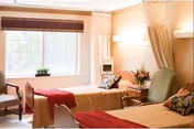A bright and cozy bedroom with two single beds, each with a red blanket and decorative pillows. There is a large window with a brown valance letting in natural light, a small table with a flower vase between the beds, and a green armchair next to one of the beds. The walls are painted a warm beige color.
