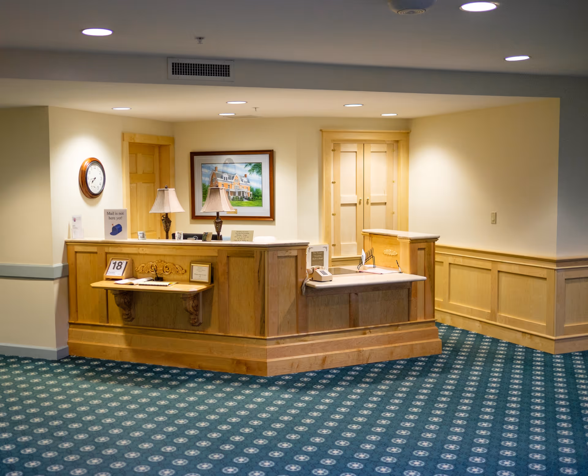 Reception desk area with wooden paneling and a green patterned carpet. The desk has two lamps, a phone, a calendar showing the 18th, and some framed signs. There is a clock on the wall, a framed picture of a house, and two wooden doors behind the desk.