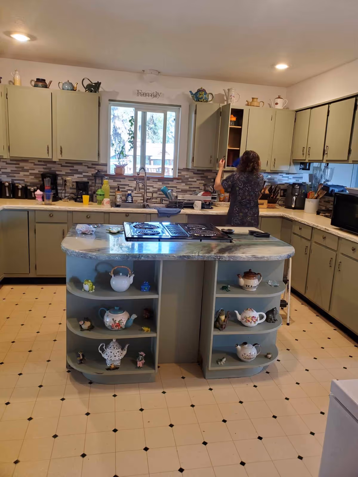 Spacious kitchen with a central island displaying teapots, pale green cabinets, and a woman reaching into an upper cabinet.
