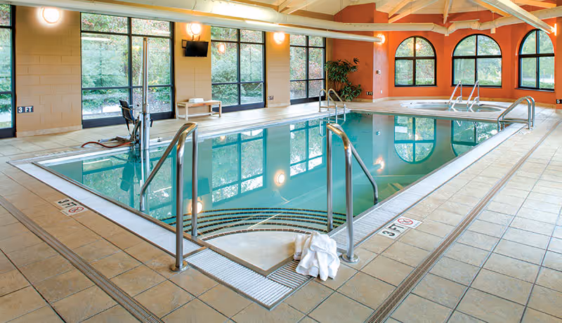Indoor swimming pool area with clear water, metal handrails, tiled floor, large windows showing greenery outside, and a hot tub in the background. A white towel is placed on the floor near the pool steps.