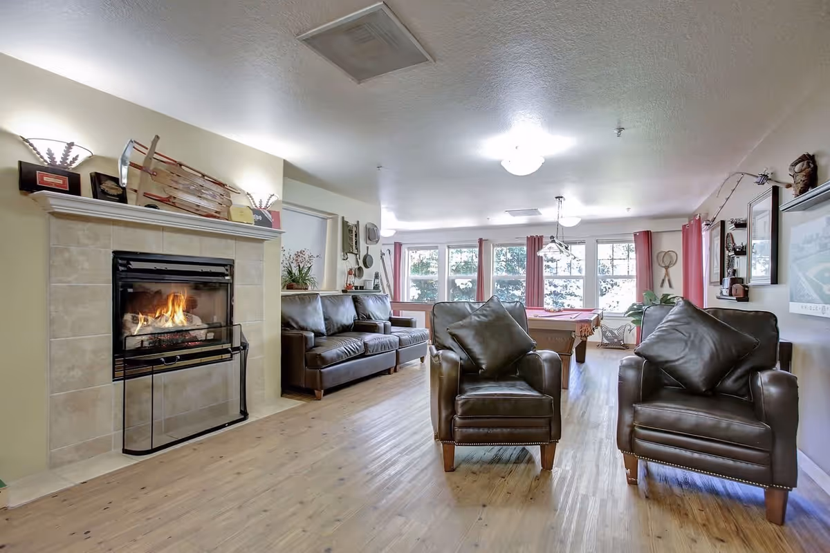 A cozy living room area with two dark leather armchairs and a matching leather sofa. There is a lit fireplace with a tiled surround on the left side. The room has wooden flooring and large windows with red curtains letting in natural light. A pool table is visible in the background along with various decorative items on the walls and shelves.