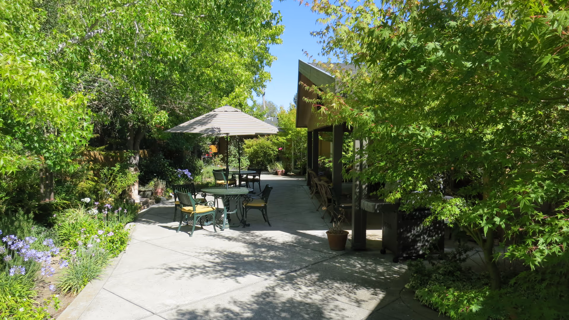 Shaded outdoor courtyard with patio tables and chairs under an umbrella surrounded by lush green trees and flowering plants.