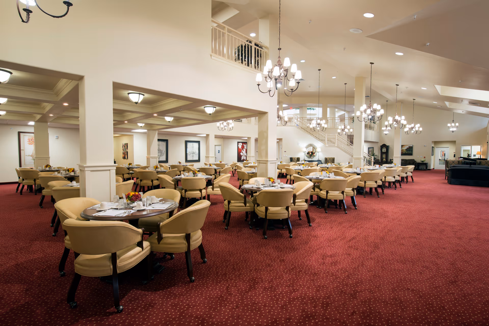 Spacious dining room with multiple round tables set with white tableware and small floral centerpieces. The room features beige upholstered chairs, red carpet with a subtle pattern, cream-colored walls, and several chandeliers hanging from the ceiling. There is a staircase and additional seating visible in the background.