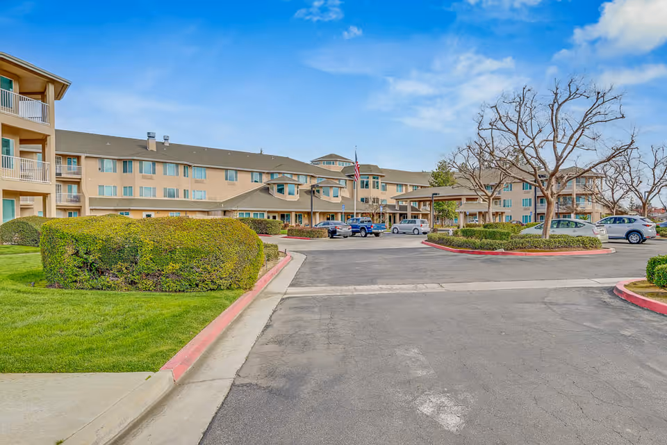 Exterior view of Solstice Senior Living at Bakersfield, showing a multi-story beige building with balconies, a parking lot with several cars, trimmed bushes, leafless trees, and a clear blue sky.
