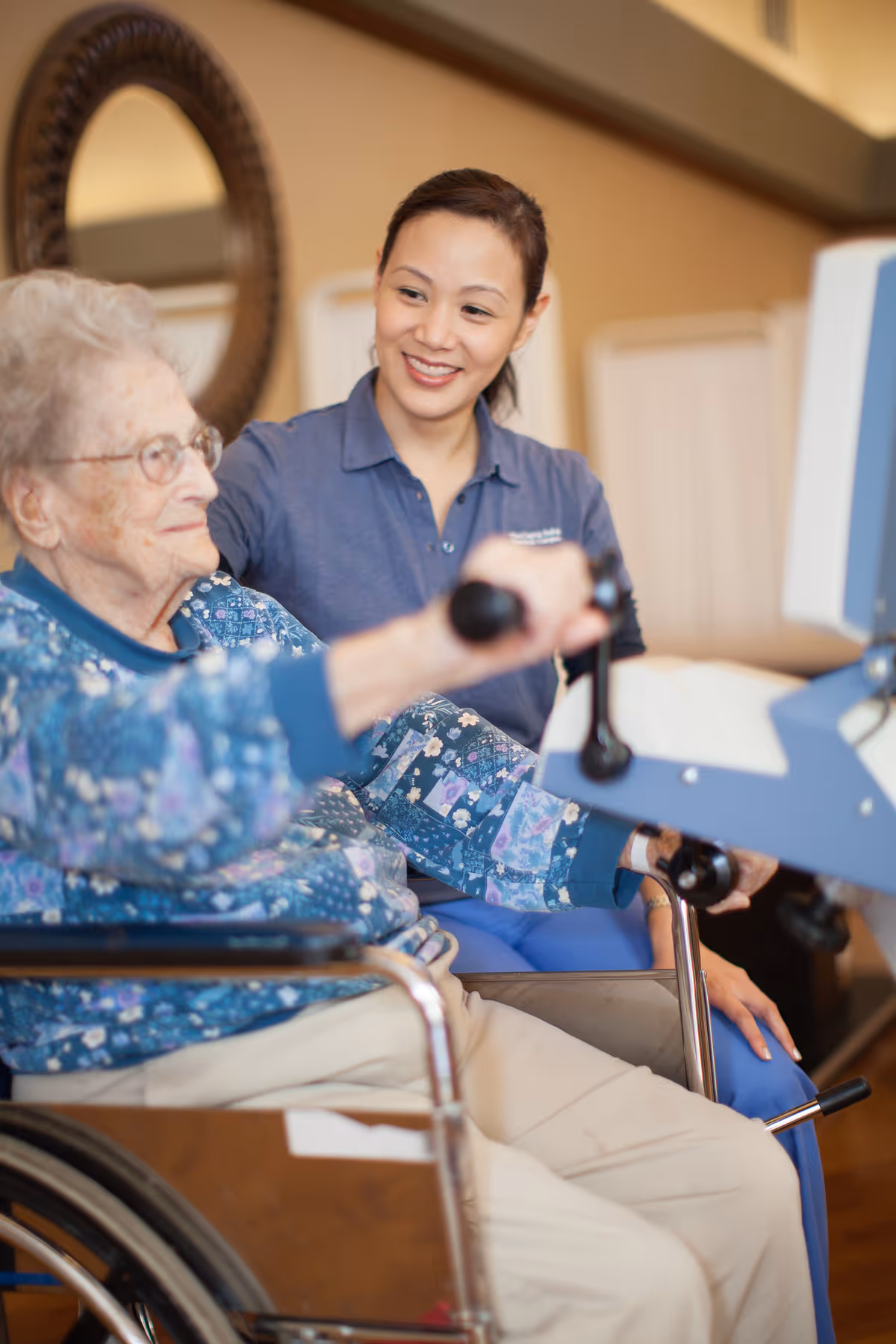 An elderly woman in a wheelchair using a piece of exercise equipment while a smiling caregiver in a blue uniform assists her in a warm indoor setting.