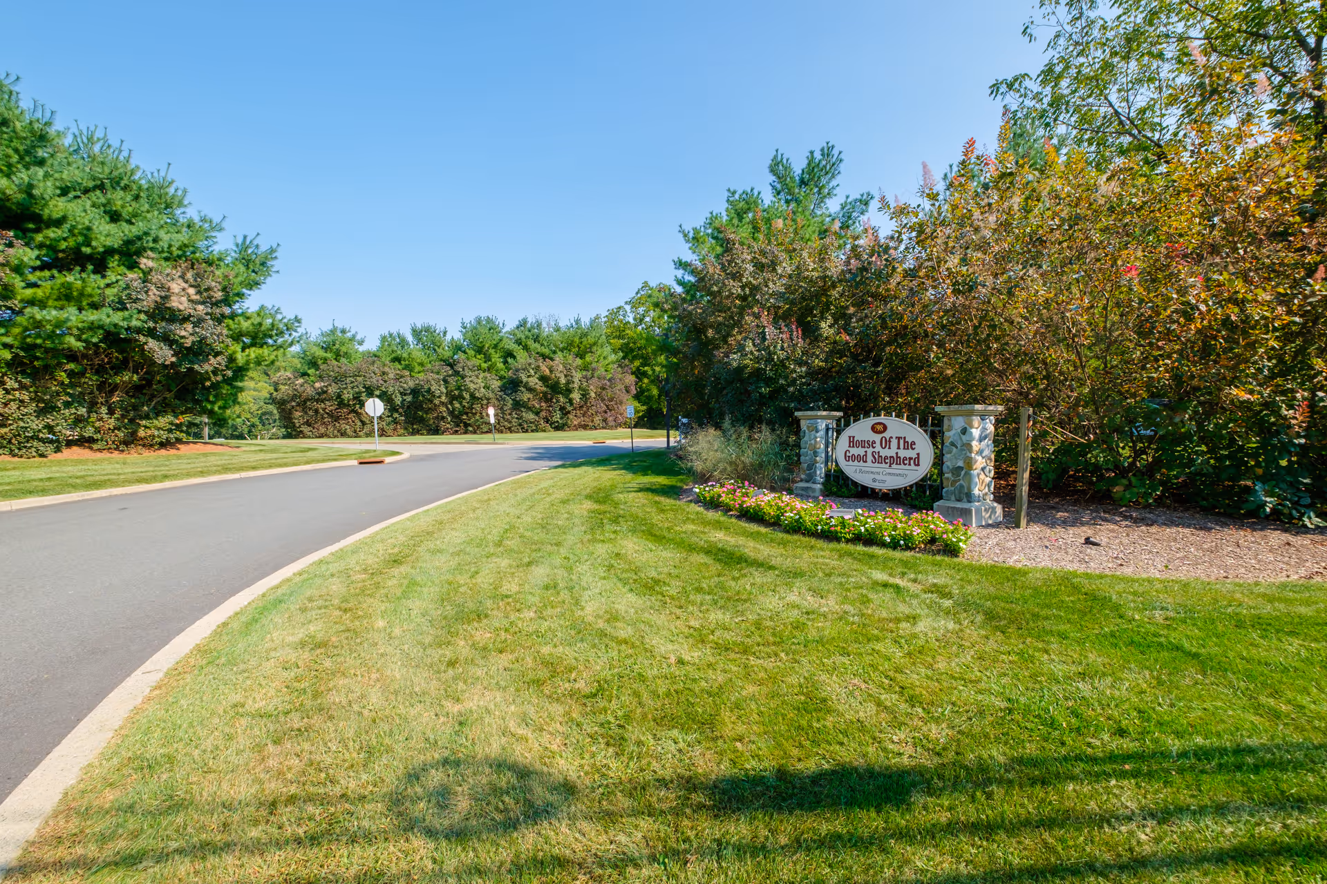 Curved driveway and well-manicured lawn leading to a stone entrance sign that reads 'House Of The Good Shepherd' surrounded by trees and flowers.