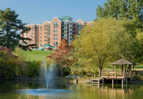Large brick senior living building beside a pond with trees and a gazebo