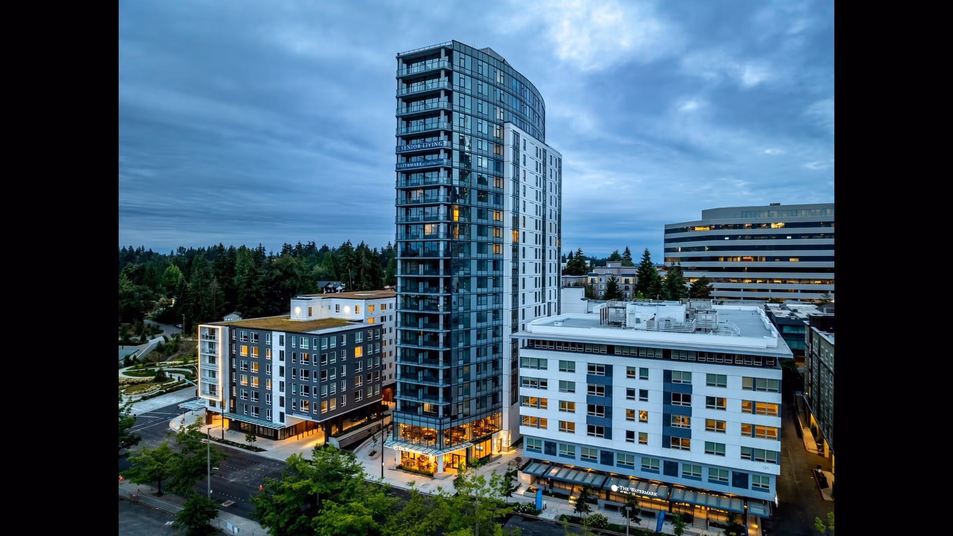 A tall modern multi-story building with glass windows and balconies, surrounded by other mid-rise buildings and greenery under a cloudy sky at dusk. The building is part of an urban area with trees and streets visible around it.