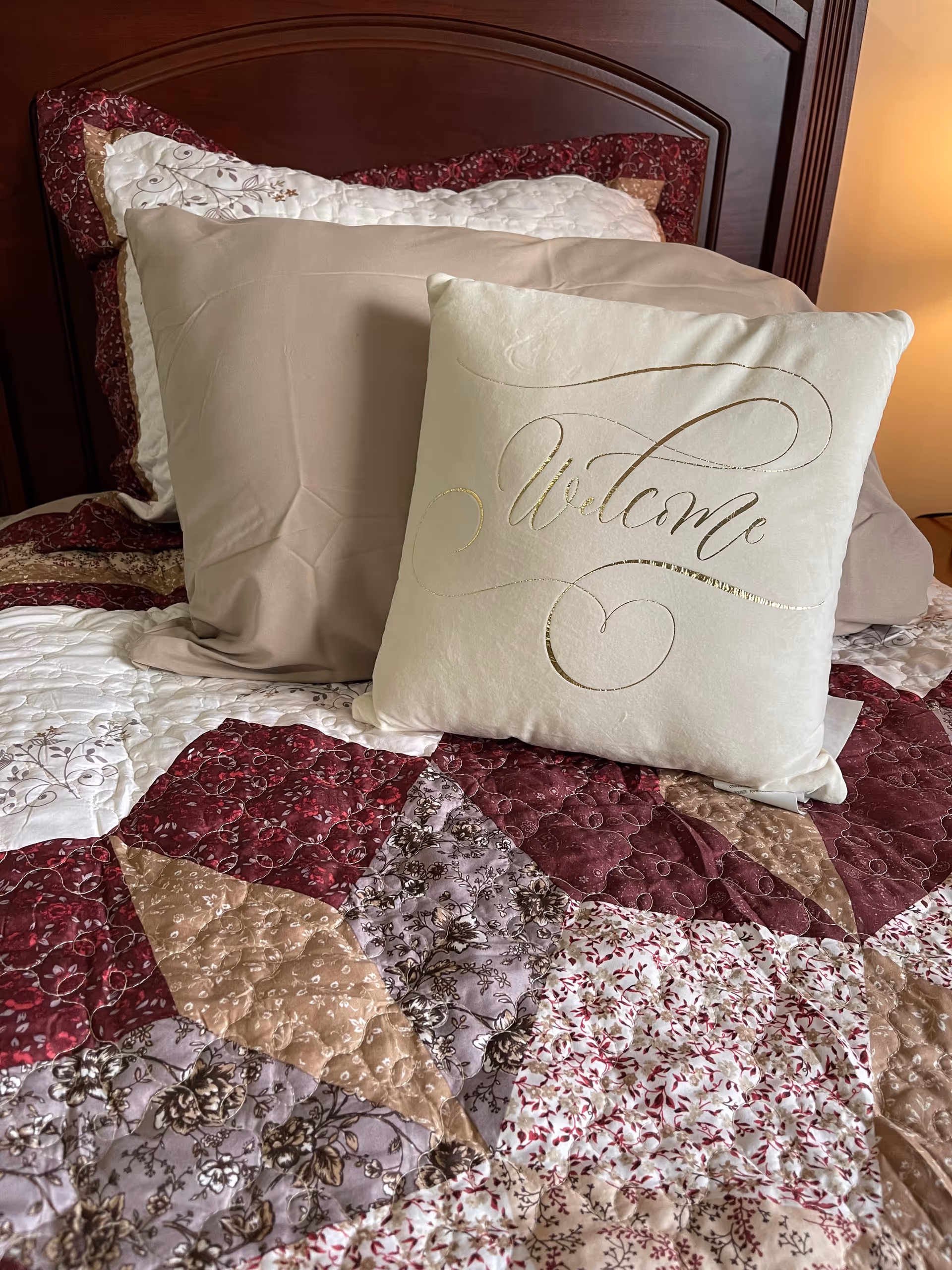 A close-up view of a bed with a wooden headboard, featuring a decorative quilt with various floral and geometric patterns in shades of burgundy, beige, and cream. On the bed are three pillows, including a prominent white pillow with the word 'Welcome' written in elegant gold script.