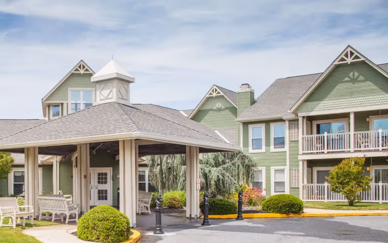 Exterior view of a senior living facility with green siding and white trim. The building has multiple windows and balconies. In front, there is a covered entrance with benches underneath and landscaped bushes around the driveway.