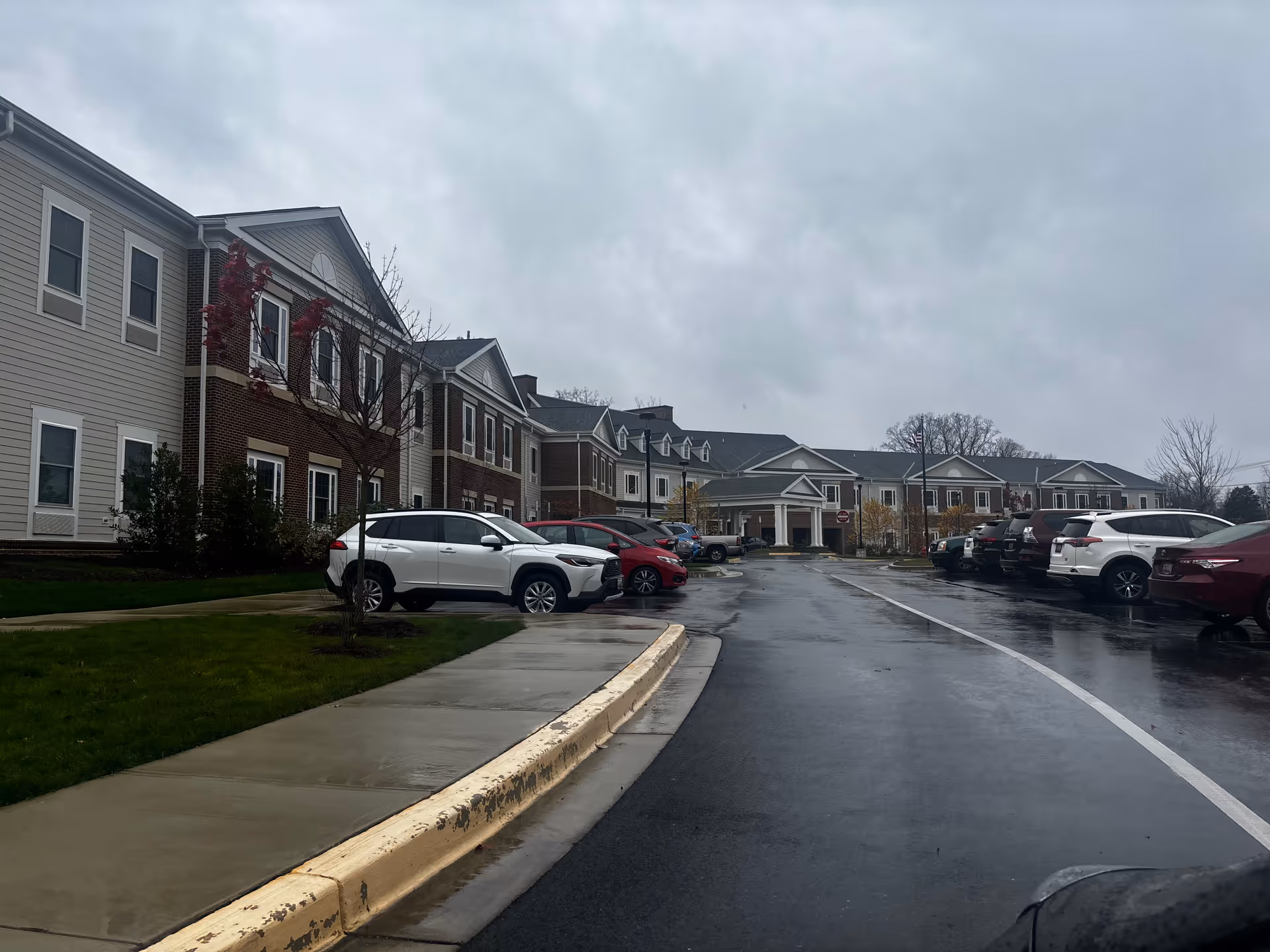Exterior view of HarborChase of Olney senior living facility on a cloudy day, showing a two-story building with brick and siding facade, a parking lot with several parked cars, and a wet driveway and sidewalk.