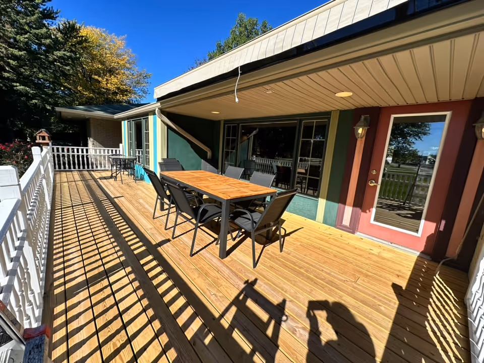Wooden outdoor deck with a dining table and chairs beside a building's exterior featuring a glass door and windows.
