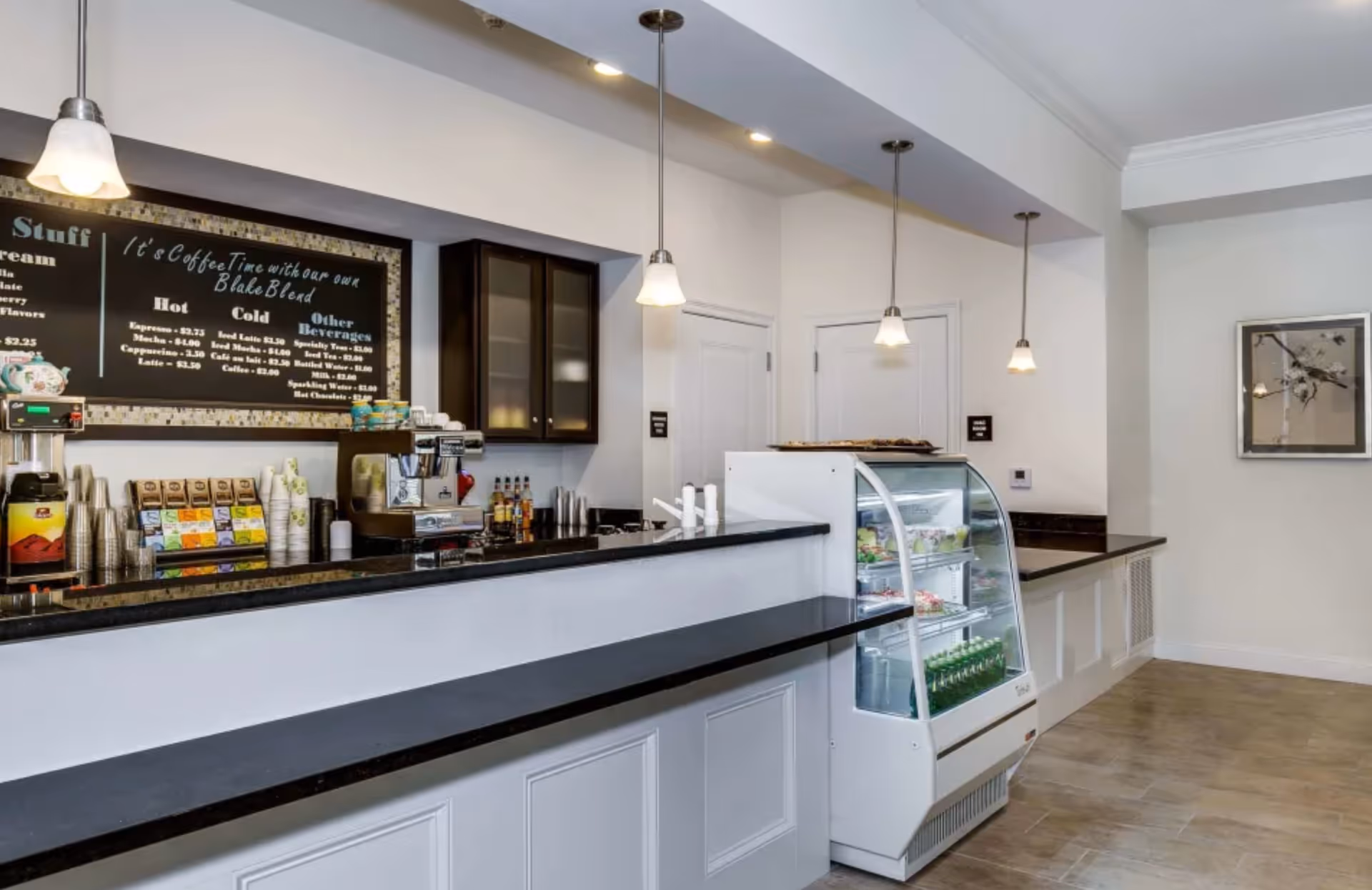 Interior view of a coffee bar area with a black countertop, white cabinetry, and hanging pendant lights. There is a menu board on the wall listing coffee and beverage options, a coffee machine, cups, and a refrigerated display case with drinks and snacks. The floor is tiled, and there is a framed picture on the wall to the right.