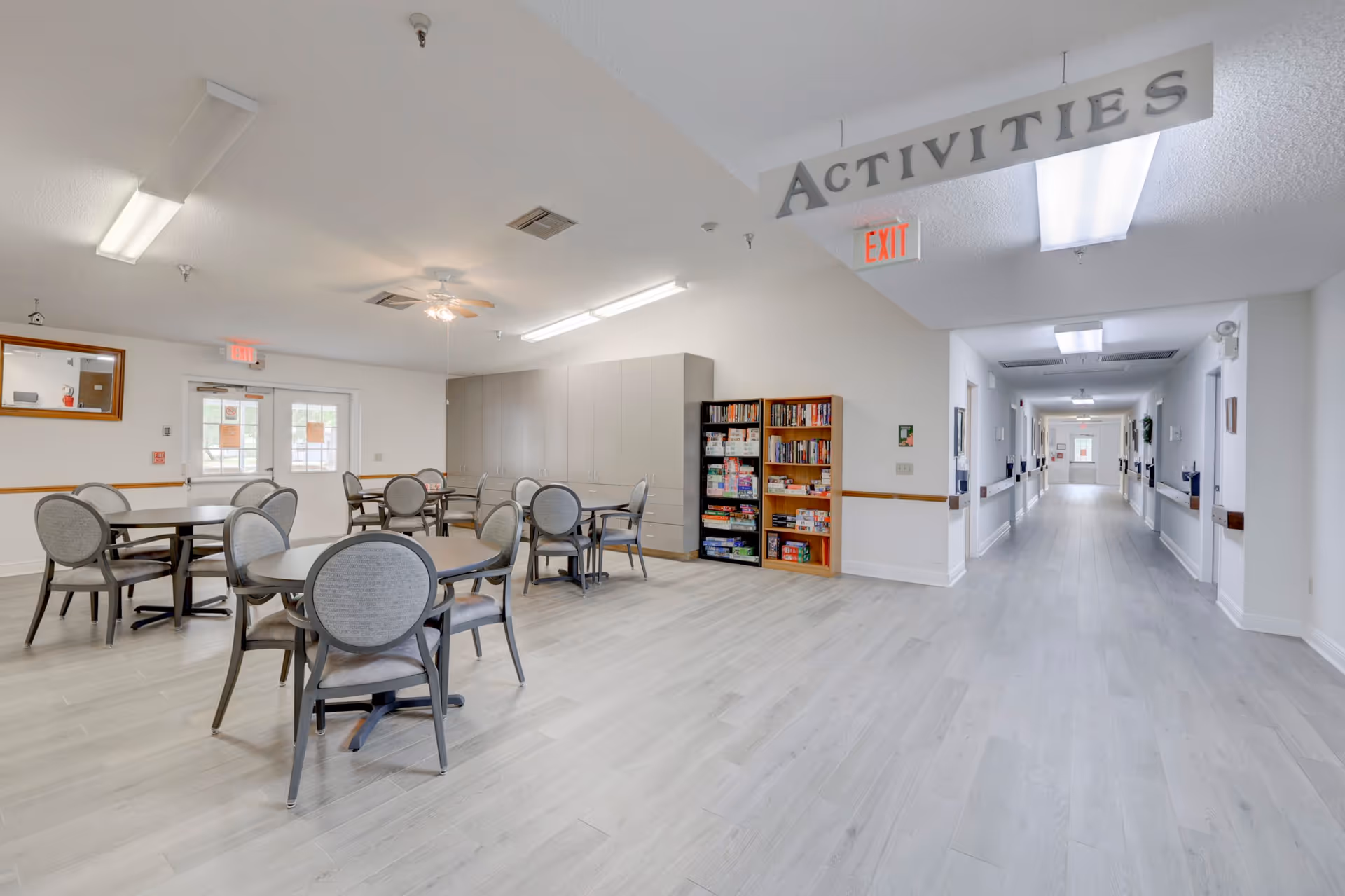 A bright and spacious activity room in a senior living facility with several round tables and chairs arranged for group activities. There is a bookshelf filled with board games and books against the wall. The room has light-colored flooring and white walls, with a hallway extending to the right. A sign hanging from the ceiling reads 'ACTIVITIES'.