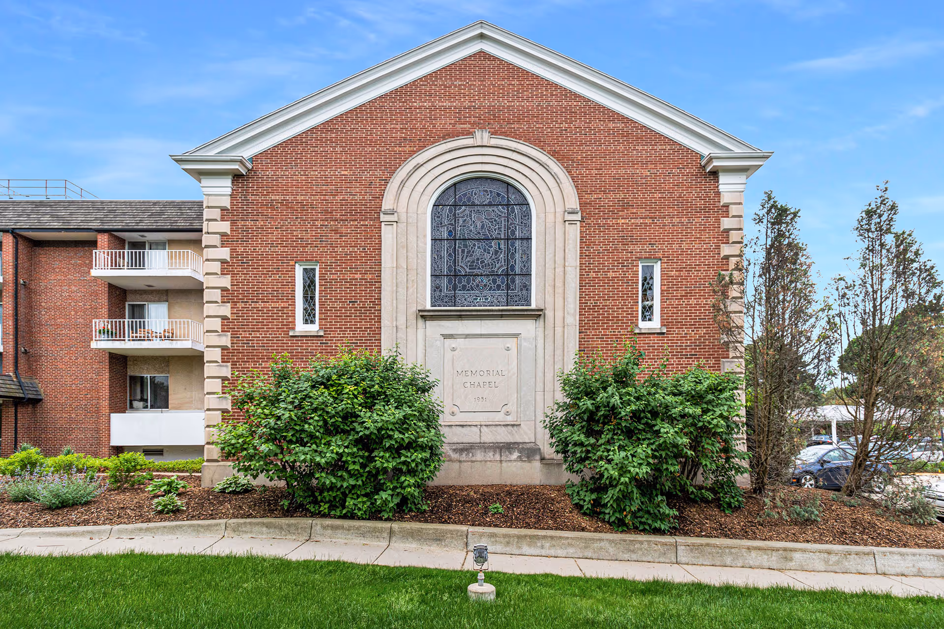 Red brick memorial chapel facade with an arched stained-glass window, flanked by shrubs and adjacent apartment balconies.