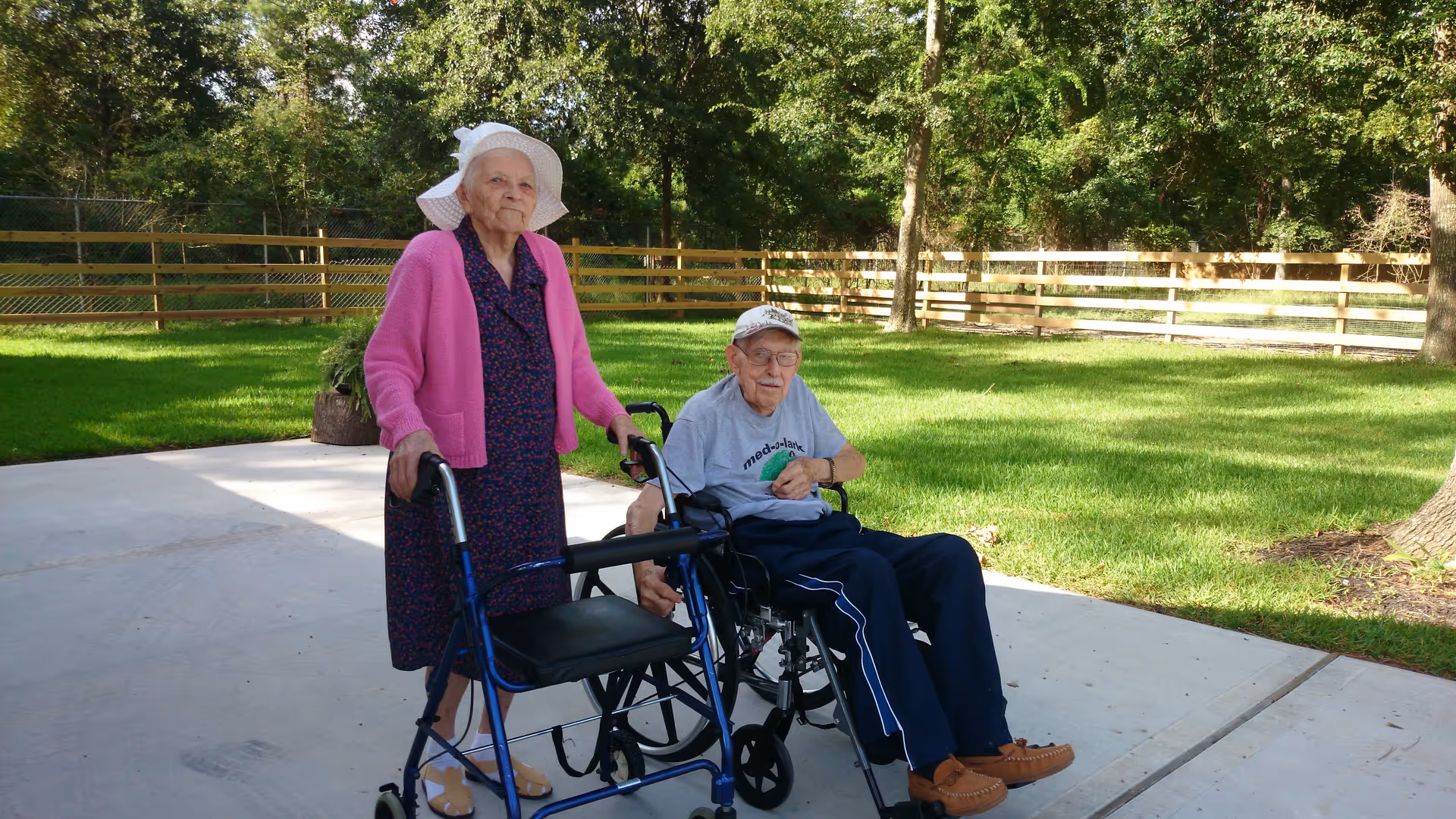 An elderly woman wearing a white sunhat, pink cardigan, and dress stands with a blue walker on a concrete patio. Next to her, an elderly man wearing a white cap, glasses, gray t-shirt, and navy blue pants sits in a wheelchair. They are outdoors with green grass, trees, and a wooden fence in the background.