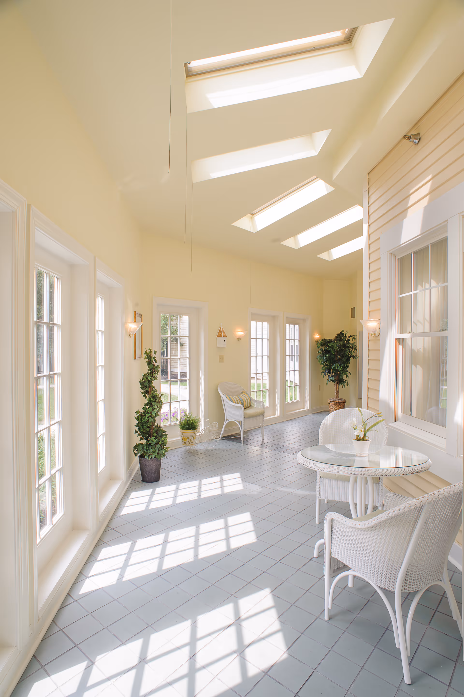 Bright sunlit indoor seating area with large windows and skylights. The space features white wicker chairs and a round glass-top table, potted plants, and light yellow walls with wall sconces. The floor is tiled in light gray, and the room has a bright, airy atmosphere.