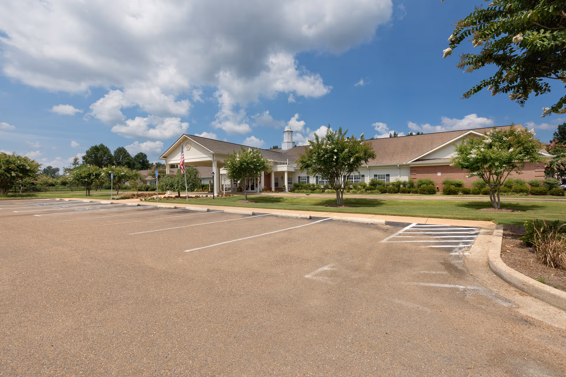 Exterior view of The Pinnacle of Oxford senior living facility showing a single-story building with a covered entrance, surrounded by green lawns and trees under a partly cloudy blue sky. An empty parking lot is in the foreground.