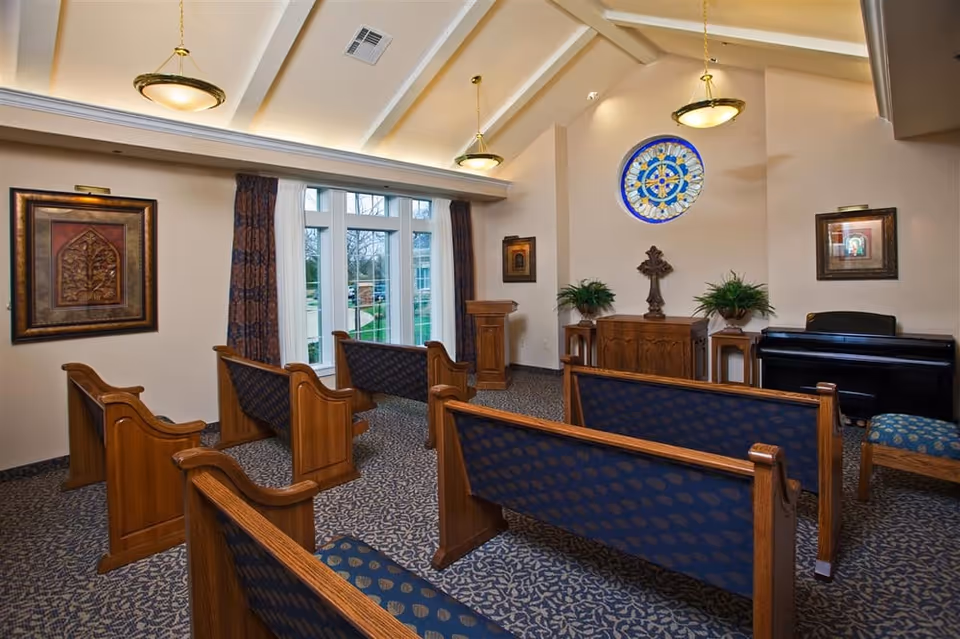 Small chapel-style meeting room with wooden pews, patterned carpet, a stained-glass window, and a piano.