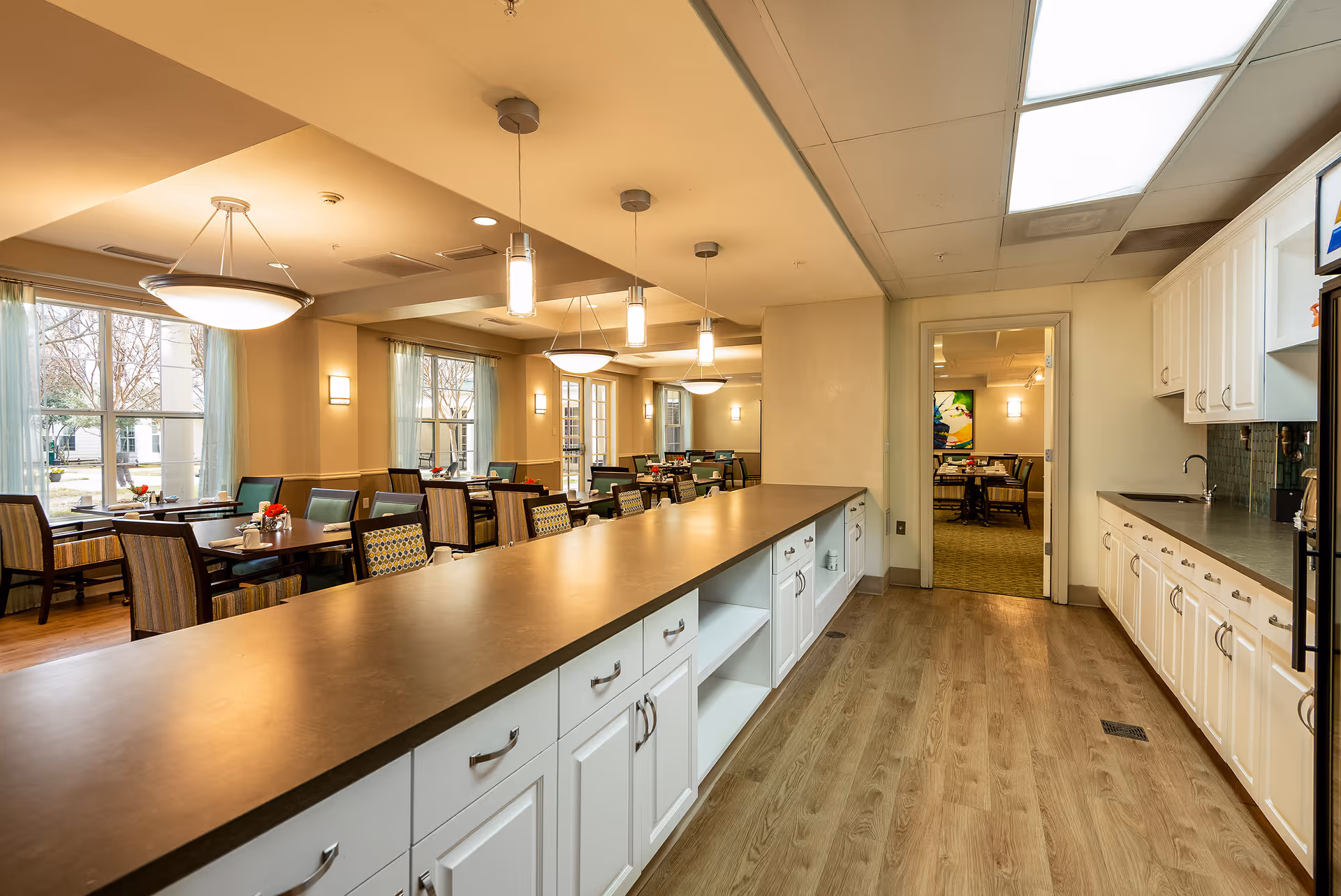 Interior view of a senior living facility dining area with multiple tables and chairs arranged near large windows with light curtains. In the foreground, there is a long countertop with white cabinets underneath and a kitchen area with white cabinetry and a sink on the right side. The room is well-lit with ceiling lights and pendant lights.