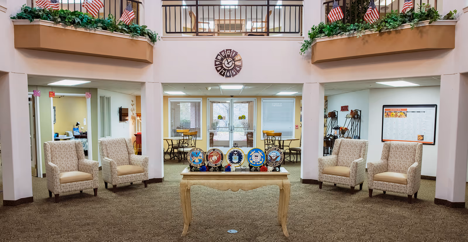 A spacious senior living community common area with four patterned armchairs arranged around a wooden table displaying military emblems. The room features a large clock on the wall, American flags, green plants on upper ledges, and a carpeted floor. In the background, there are tables and chairs near glass doors leading outside.