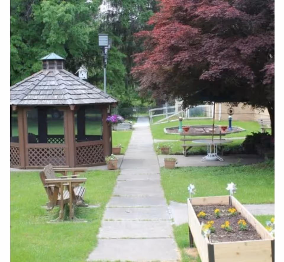 A paved walkway through a grassy courtyard with a wooden gazebo, benches, planter boxes and a large tree.
