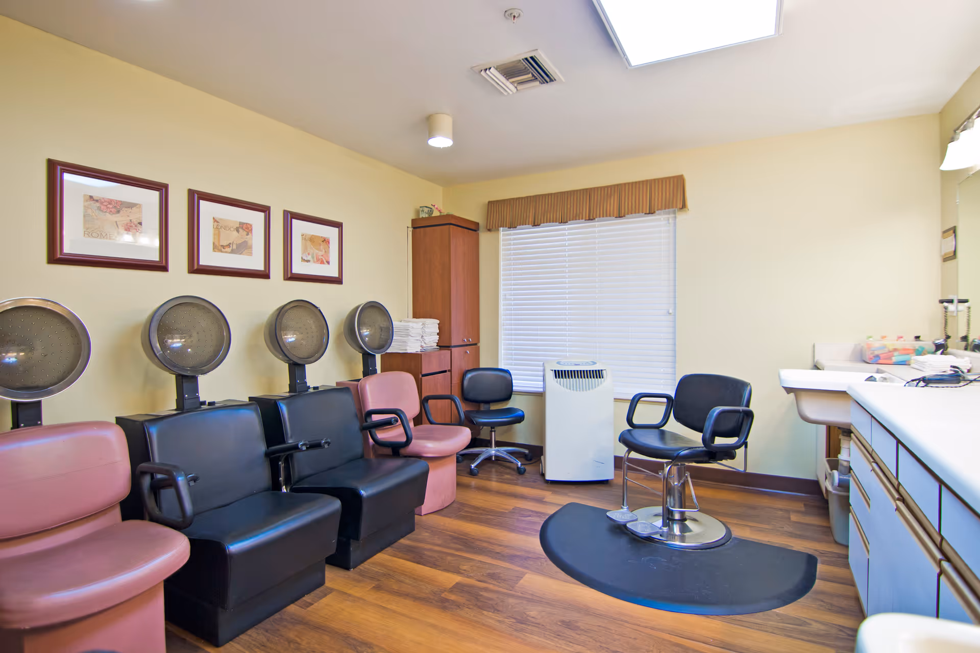 Interior view of a salon room with four black and two pink salon chairs, each with hair dryers above them. There is a window with blinds and a brown valance, a portable air conditioner, a swivel salon chair on a black mat, and a counter with a sink and various supplies. The walls are painted light yellow and the floor is wooden.