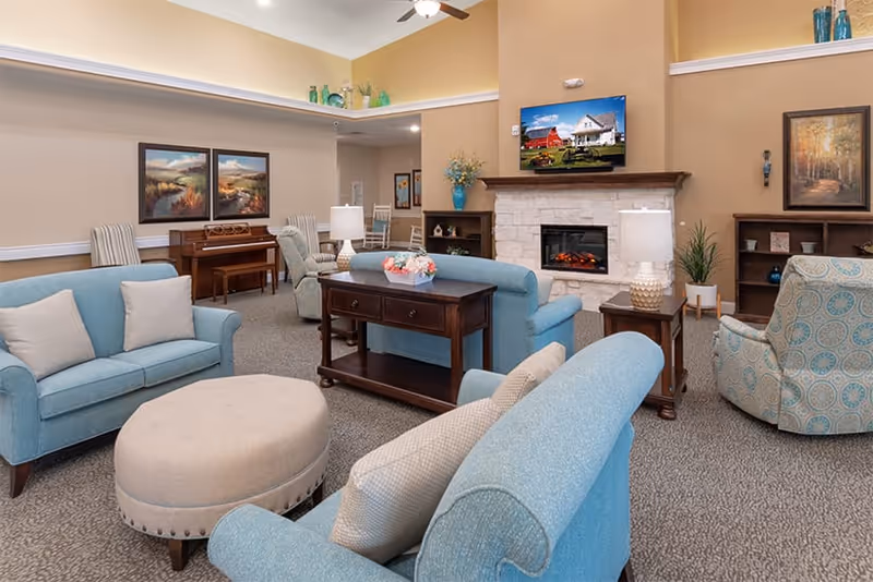 A cozy living room in an assisted living facility featuring light blue upholstered sofas and armchairs arranged around a round beige ottoman. The room has a stone fireplace with a TV mounted above it, wooden side tables with lamps, a piano against the wall, and decorative paintings and vases. The walls are painted in warm beige and yellow tones, and the carpet is gray.