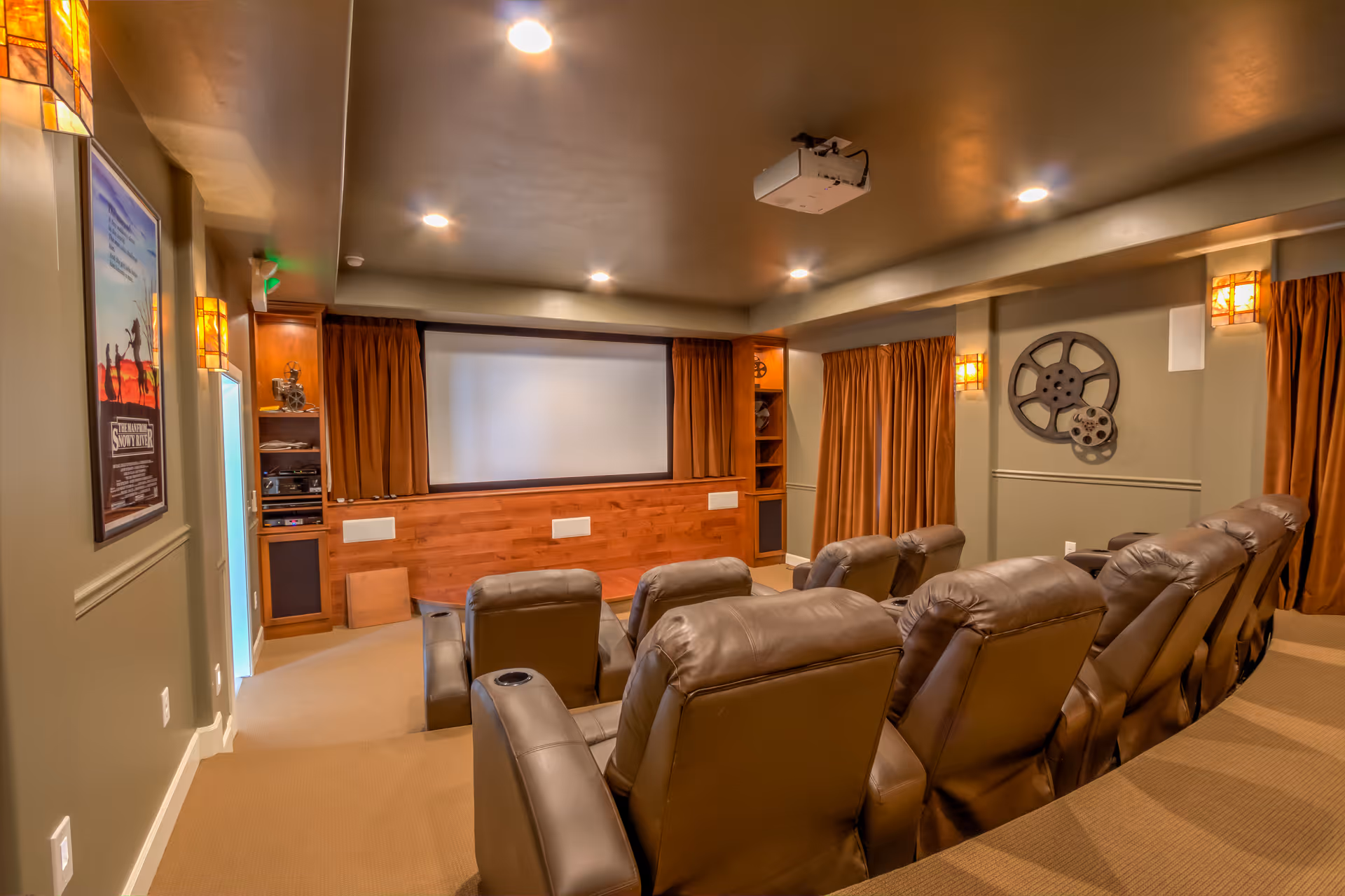 A cozy home theater room with two rows of brown leather recliner chairs facing a large white projection screen. The room has warm lighting with wall sconces, brown curtains covering the windows, and movie-themed decorations including film reels on the wall and a framed movie poster. A ceiling-mounted projector is visible above the seating area.