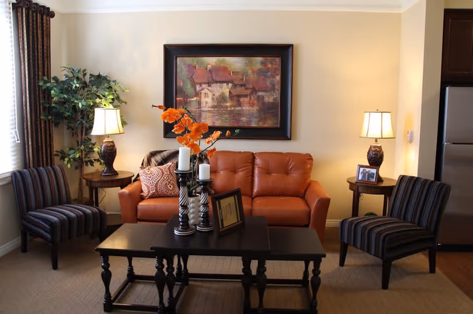 A cozy living room with a brown leather sofa adorned with patterned pillows, two striped armchairs, a dark wooden coffee table with decorative candles and a framed picture, two side tables each with a lamp, a potted plant in the corner, and a large framed painting of houses on the wall behind the sofa.