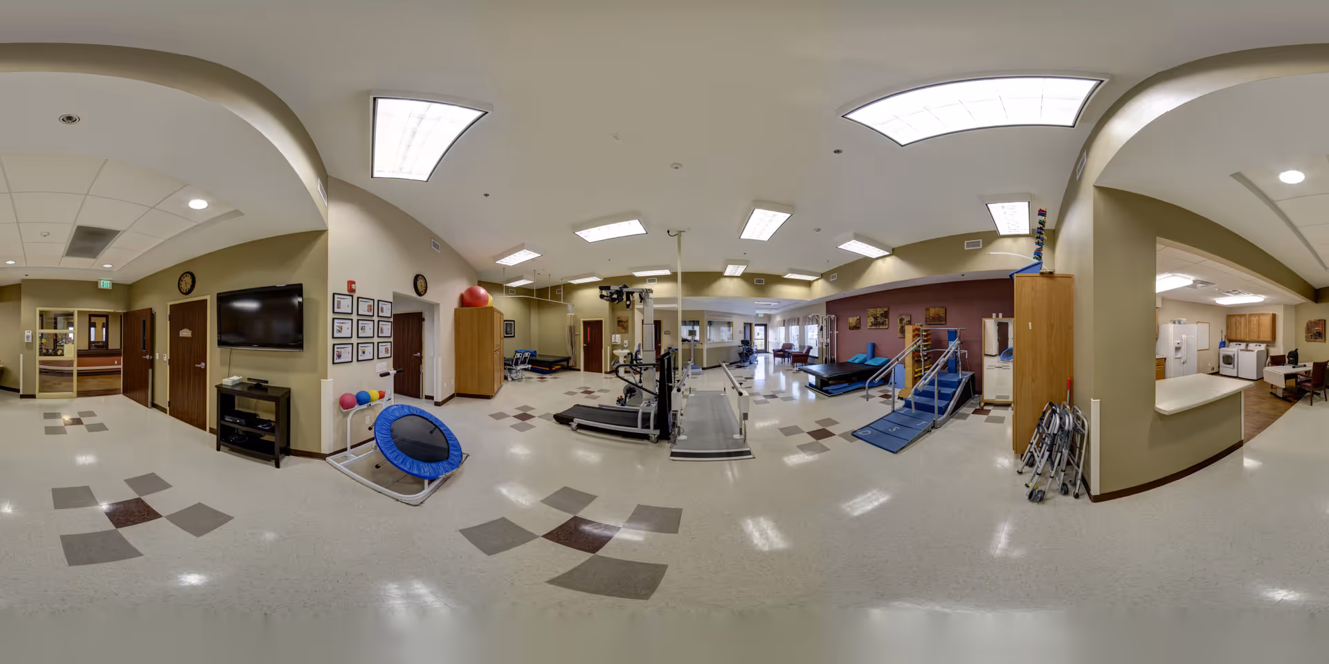 A spacious physical therapy and rehabilitation room in Sunnyside Healthcare Center featuring exercise equipment including a treadmill, parallel bars, a small trampoline, therapy balls, and a set of stairs with handrails. The room has beige walls, tiled flooring with a pattern, fluorescent ceiling lights, and several doors leading to other areas. There is also a TV mounted on the wall and a small seating area in the background.