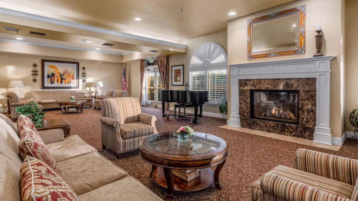 A cozy living room area in North Shore Place featuring a fireplace with a marble surround and a large mirror above it. The room has multiple seating options including striped armchairs and beige sofas with patterned cushions. A round glass-top coffee table with a small flower arrangement is in the center. There is a black grand piano near a window with white shutters and an American flag in the corner. The room is warmly lit with table lamps and wall sconces, and has carpeted flooring with a patterned design.