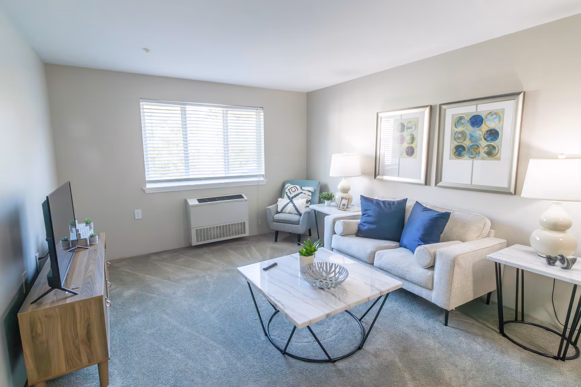 A bright living room with a beige sofa adorned with two blue pillows, a gray armchair with a patterned pillow, a marble-top coffee table with a decorative bowl and a remote control, two side tables each with a white lamp, two framed abstract artworks on the wall, a wooden TV stand with a flat-screen TV, and a window with blinds letting in natural light.