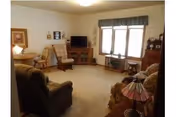 Carpeted living room with upholstered chairs and a sofa, a TV on a wooden stand, lamps, and a window with a valance.