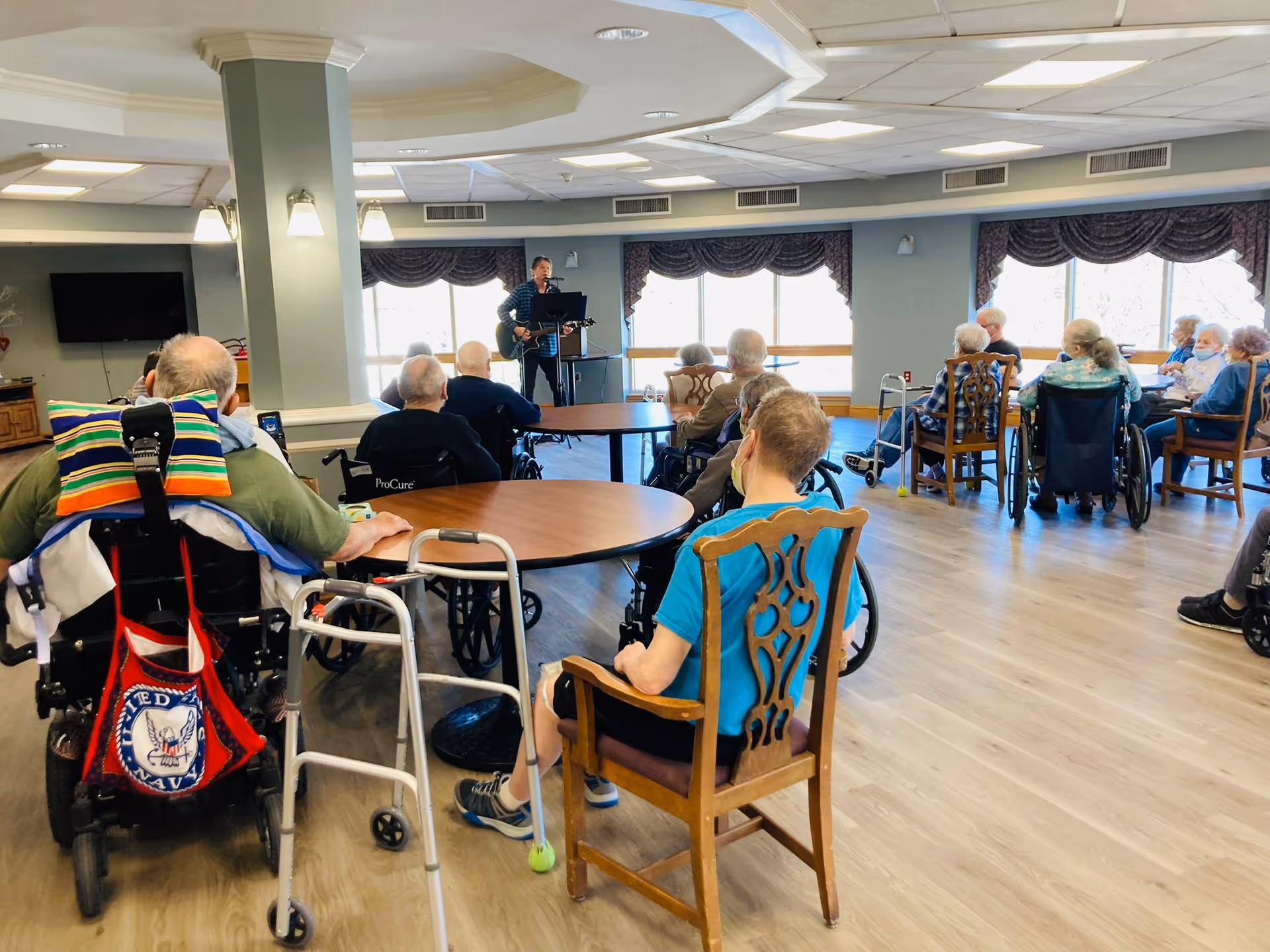 A group of elderly people, some in wheelchairs, seated around tables in a common room watching a man playing guitar and singing near the windows.