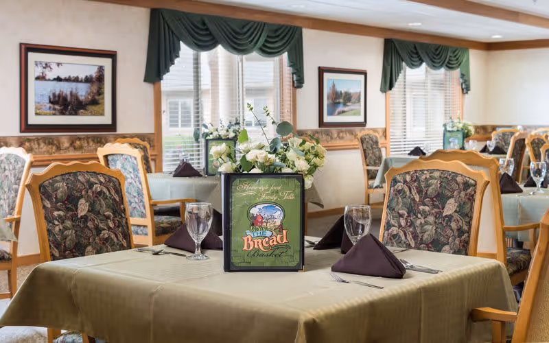 A dining room with tables covered in beige tablecloths, each set with folded dark napkins, water glasses, and silverware. The chairs have floral patterned upholstery. A menu titled 'The Bread Basket' and a floral centerpiece are placed on the table in the foreground. The room has large windows with green valances and framed landscape pictures on the walls.