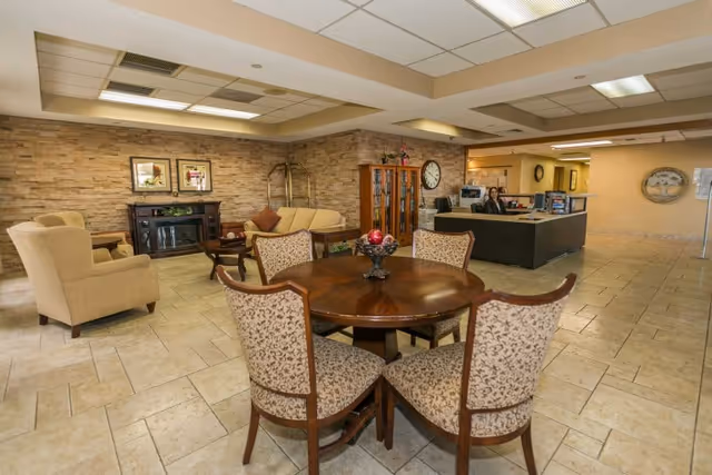 A spacious common area in a senior living facility featuring a round wooden table with four upholstered chairs in the foreground. In the background, there is a seating area with beige armchairs and a sofa, a wooden cabinet with glass doors, a clock on the wall, and a reception desk with a staff member seated behind it. The walls have a stone-like texture, and the floor is tiled.