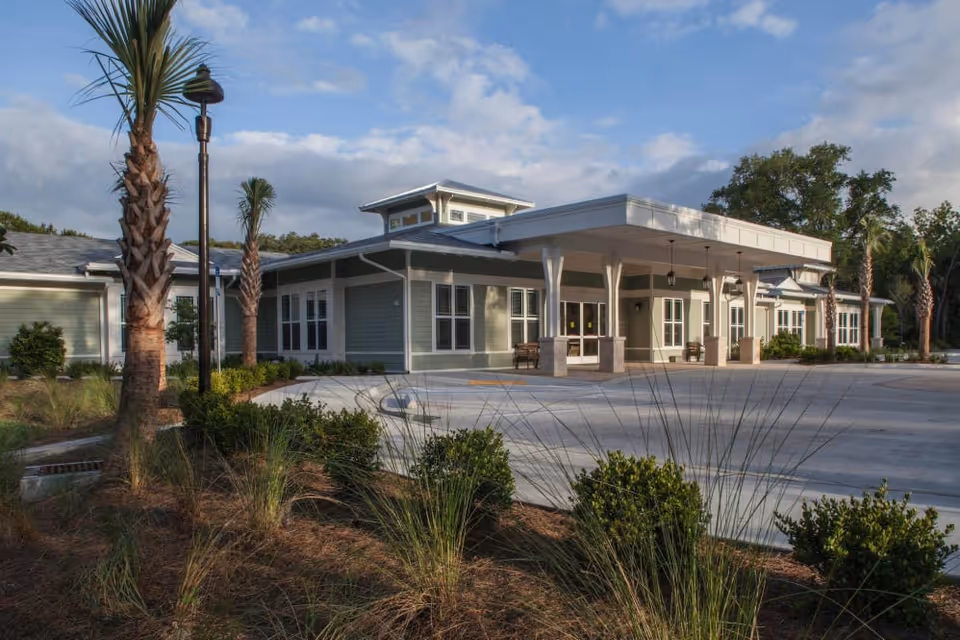Exterior view of a single-story building with a covered entrance, surrounded by landscaped greenery including palm trees and bushes under a partly cloudy sky.