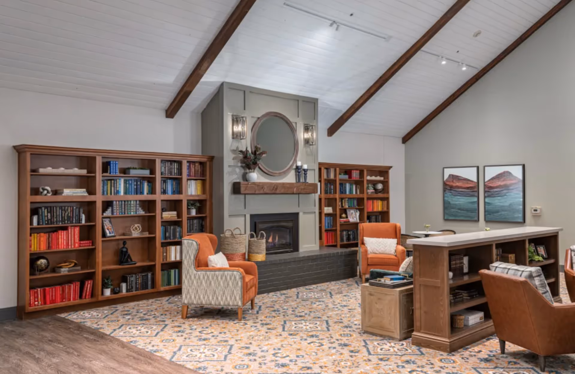A cozy senior living common area with a high white ceiling featuring wooden beams, a fireplace with a round mirror above it, flanked by two wooden bookshelves filled with books and decorative items. The room has patterned carpet, two orange armchairs with pillows, a wooden bookshelf divider, and two framed landscape paintings on the wall.