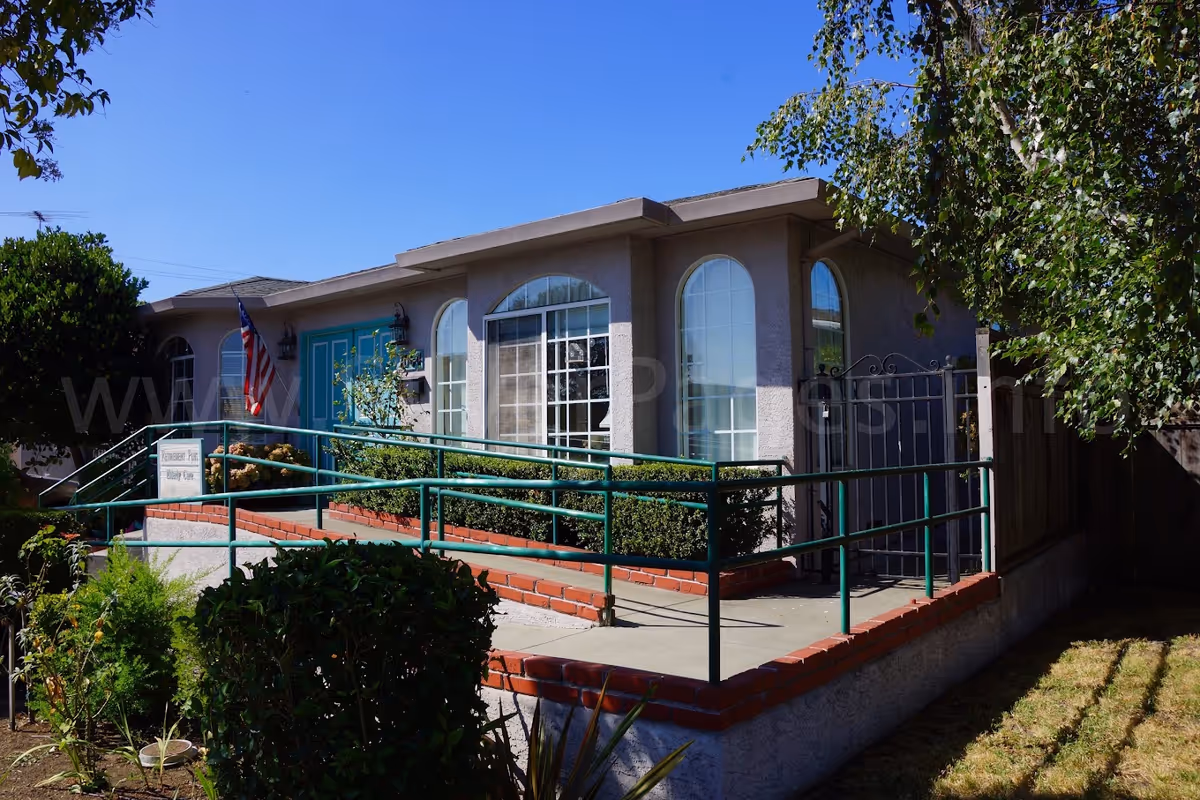 Front entrance of a single-story care home with a wheelchair ramp, teal doors, arched windows, and an American flag.
