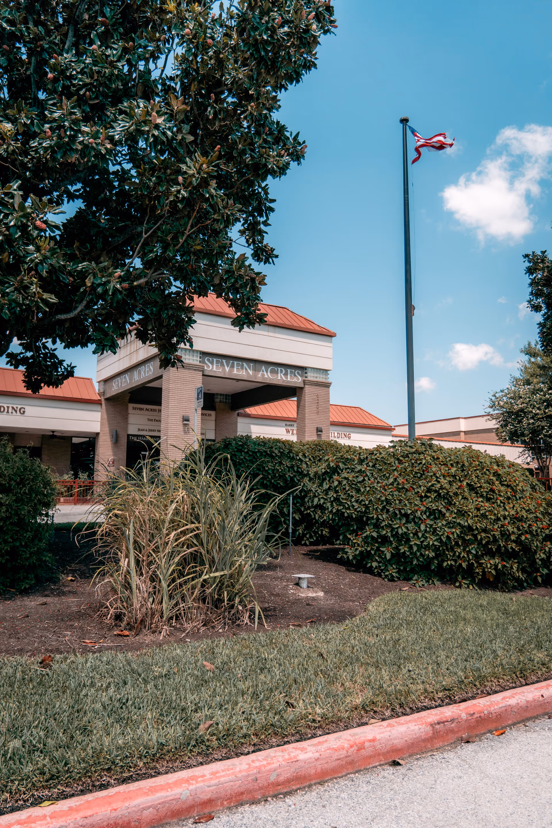 Exterior view of the Seven Acres Jewish Senior Care Services building with a flagpole flying an American flag, surrounded by green bushes and trees under a clear blue sky.
